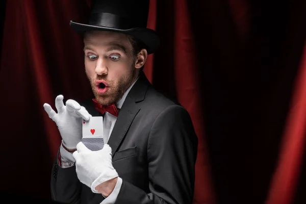 Magicien choqué tenant des cartes à jouer dans le cirque avec des rideaux rouges — Photo de stock