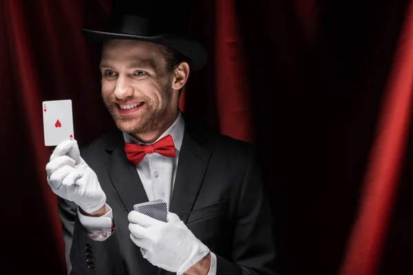 Cheerful magician holding playing cards in circus with red curtains — Stock Photo