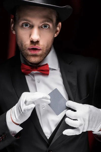 Surprised magician holding playing card in circus with red curtains — Stock Photo