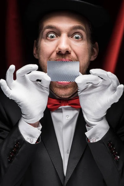 Scary magician showing trick with playing card in circus with red curtains — Stock Photo