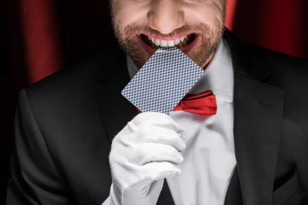Cropped view of smiling magician holding playing card in teeth in circus with red curtains — Stock Photo