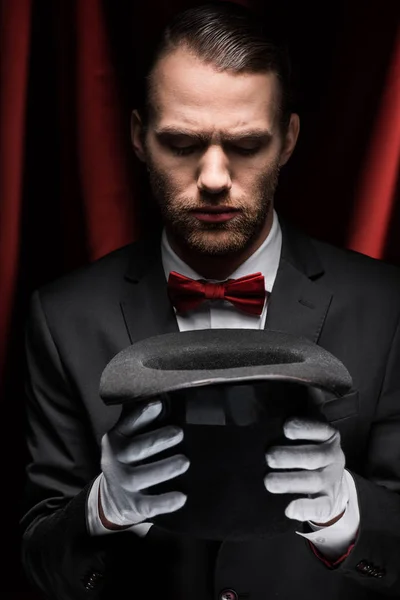 Focused magician in suit looking at hat in circus with red curtains — Stock Photo