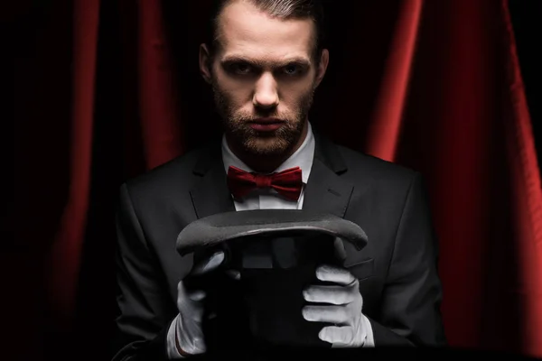 Concentrated magician in suit holding hat in circus with red curtains — Stock Photo