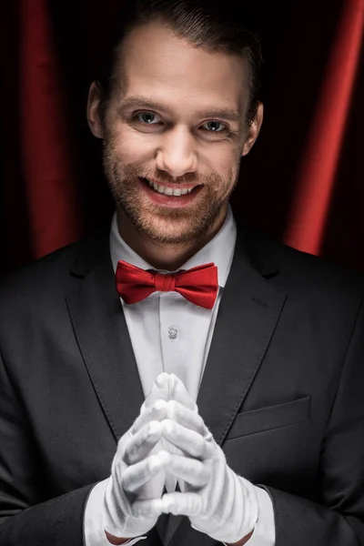 Cavalheiro sorridente de terno, gravata borboleta e luvas em circo com cortinas vermelhas — Fotografia de Stock