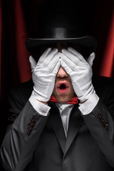 Scared magician closing eyes with hands in gloves in circus with red curtains — Stock Photo