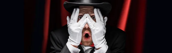 Panoramic shot of frightened magician closing eyes with hands in gloves in circus with red curtains — Stock Photo