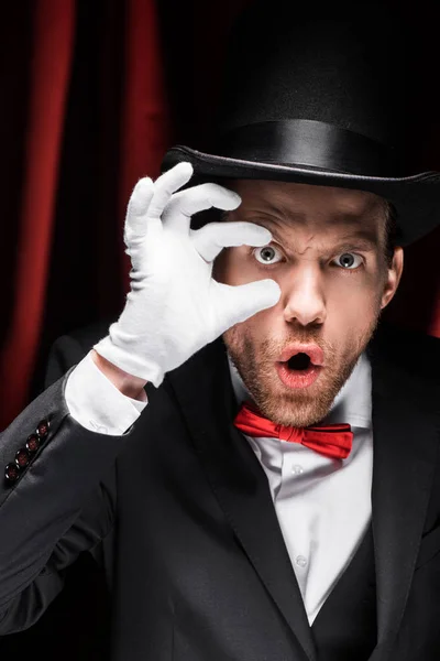 Schockierter Zauberer öffnet Augen mit Fingern im Zirkus mit roten Vorhängen — Stockfoto