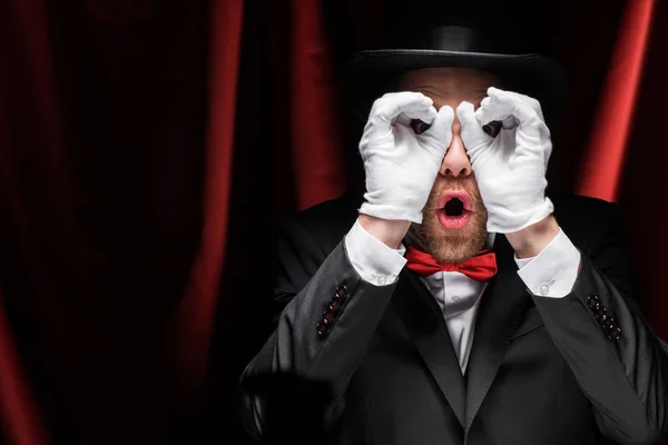 Shocked magician making glasses from hands in circus with red curtains — Stock Photo