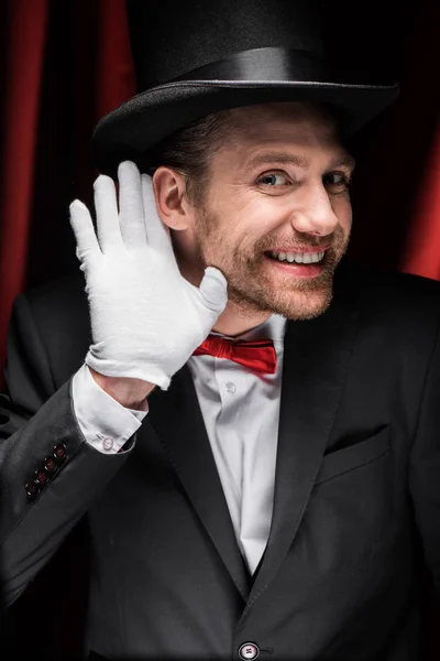 Mago sonriente haciendo gestos y escuchando en circo con cortinas rojas — Stock Photo