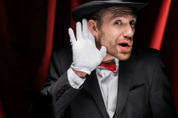 Confused magician gesturing and listening in circus with red curtains — Stock Photo