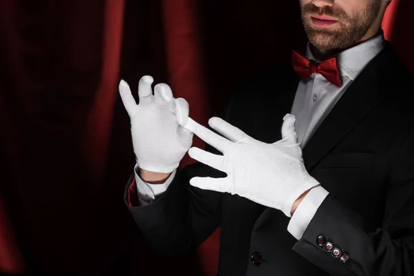 Cropped view of magician taking off white gloves in circus with red curtains — Stock Photo