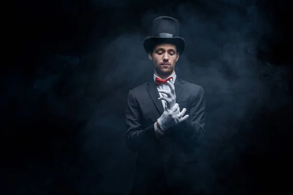 Magician in suit and hat wearing gloves in dark smoky room — Stock Photo