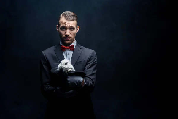 Professional young magician holding white rabbit in hat, isolated on black — Stock Photo