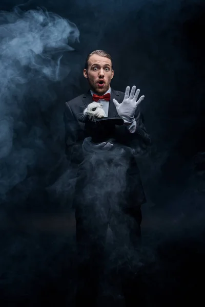 Emotional magician in suit holding hat with white rabbit, dark room with smoke — Stock Photo