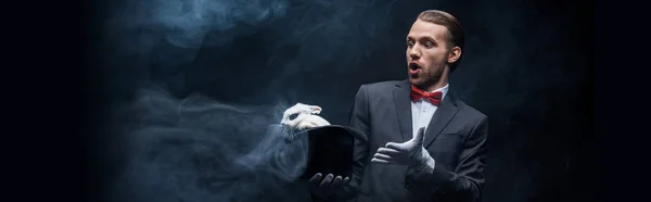 Panoramic shot of shocked magician in suit holding hat with white rabbit, dark room with smoke — Stock Photo