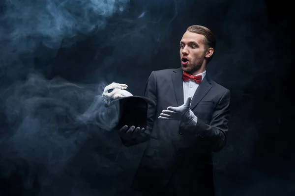 Mágico surpreso em terno mostrando truque com coelho branco em chapéu, sala escura com fumaça — Fotografia de Stock