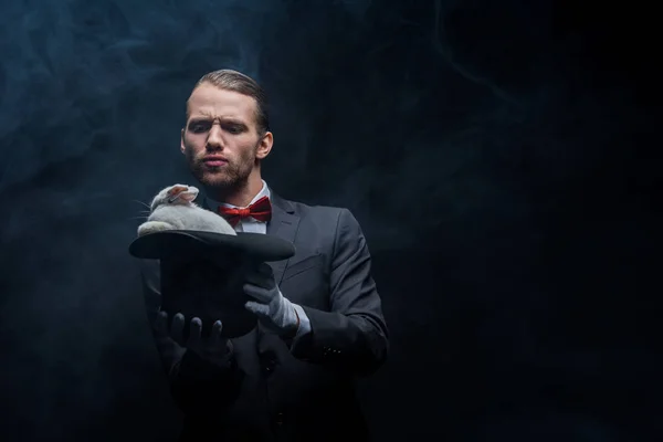 Emotional magician in suit showing trick with white rabbit in hat, dark room with smoke — Stock Photo