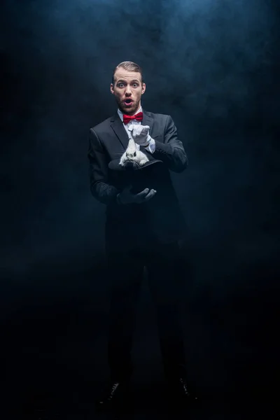 Aufgeregter Zauberer im Anzug zeigt Trick mit weißem Kaninchen im Hut, dunkler Raum mit Rauch — Stockfoto