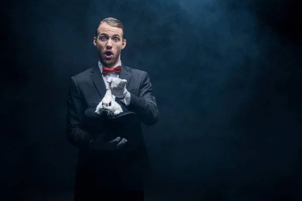 Magicien choqué avec bouche ouverte montrant tour avec lapin blanc dans le chapeau, chambre sombre avec de la fumée — Photo de stock