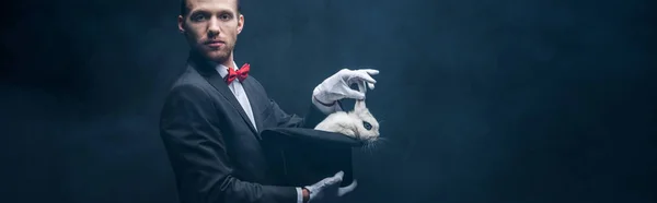 Panoramic shot of young magician in suit showing trick with white rabbit in hat, dark room with smoke — Stock Photo