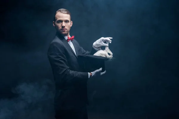 Professional magician in suit showing trick with white rabbit in hat, dark room with smoke — Stock Photo