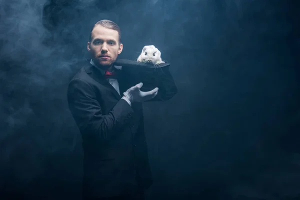 Mago profesional en traje mostrando truco con conejo blanco en sombrero, cuarto oscuro con humo - foto de stock