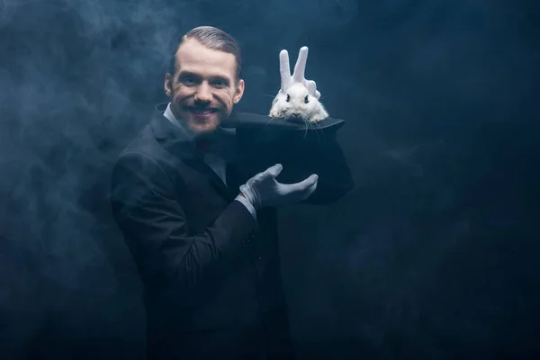 Positive magician in suit showing trick with white rabbit in hat, dark room with smoke — Stock Photo