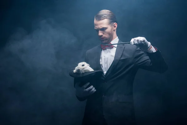 Mago serio en traje mostrando truco con varita y conejo blanco en sombrero, cuarto oscuro con humo - foto de stock