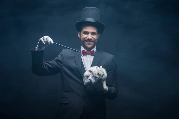 Cheerful magician in suit showing trick with wand and white rabbit in hat, dark room with smoke — Stock Photo