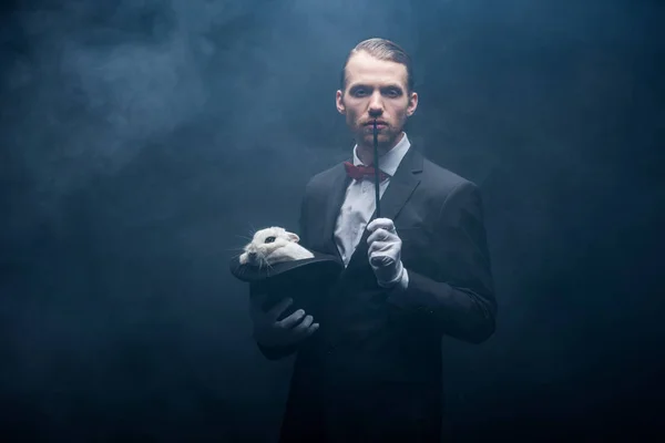 Magicien en costume montrant tour avec baguette et lapin blanc dans le chapeau, chambre sombre avec fumée — Photo de stock
