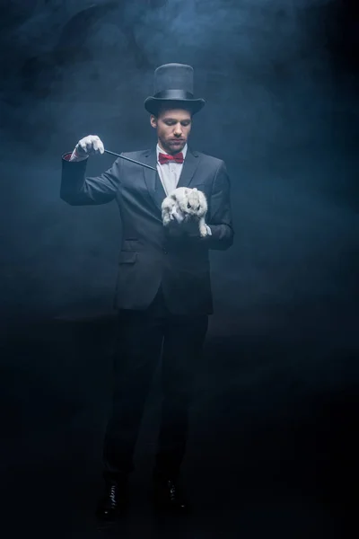 Profesional joven mago en traje y sombrero mostrando truco con varita y conejo blanco, cuarto oscuro con humo - foto de stock