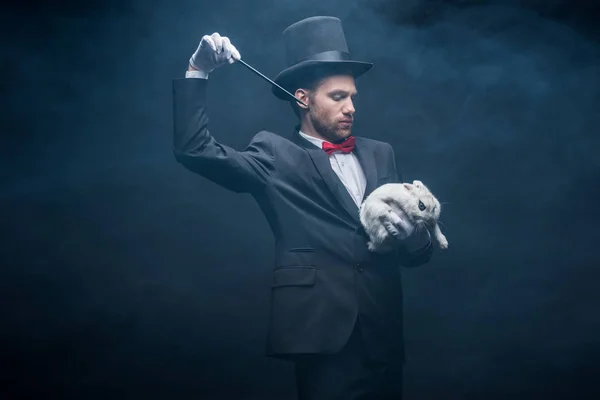 Emotional magician in suit and hat showing trick with wand and white rabbit, dark room with smoke — Stock Photo