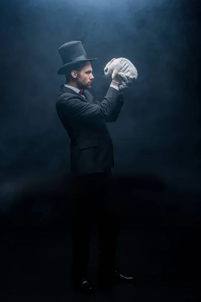 Focused magician in suit and hat holding white bunny, dark room with smoke — Stock Photo