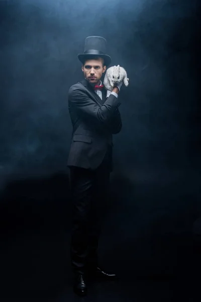 Magicien sérieux en costume et chapeau tenant lapin blanc, chambre sombre avec fumée — Photo de stock