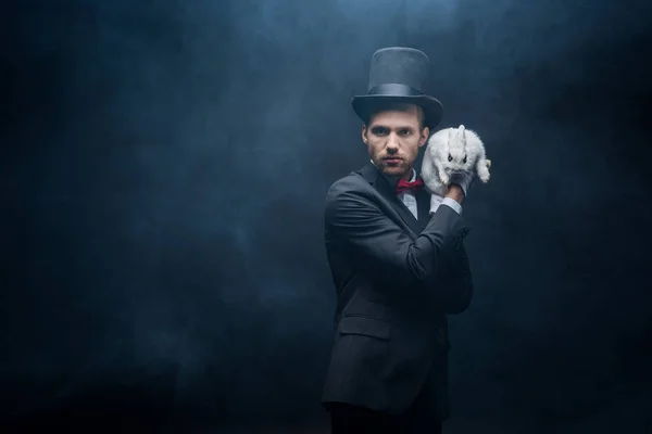 Magicien professionnel en costume et chapeau tenant lapin blanc, chambre sombre avec fumée — Photo de stock