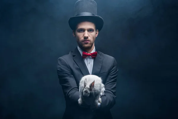 Mago confiado en traje y sombrero sosteniendo conejo blanco, habitación oscura con humo - foto de stock