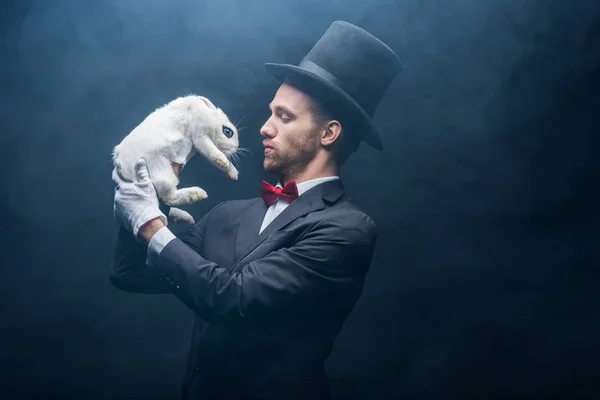 Mago profesional en traje y sombrero mirando conejo blanco, cuarto oscuro con humo - foto de stock