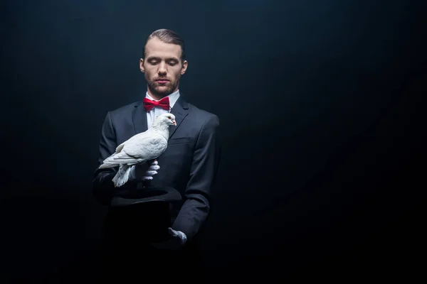 Young magician showing trick with dove and hat in dark room with smoke — Stock Photo