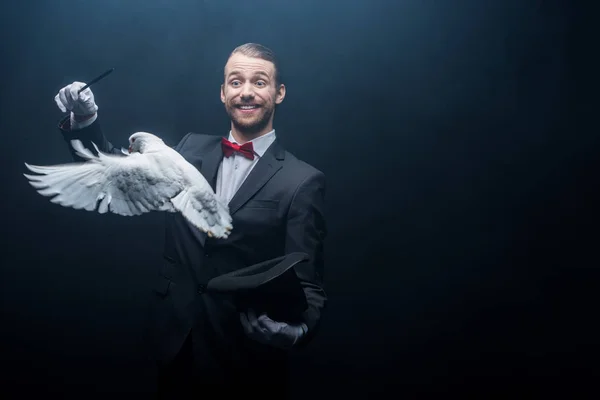 Lächelnder Zauberer zeigt Trick mit Taube, Zauberstab und Hut in dunklem Raum mit Rauch — Stockfoto