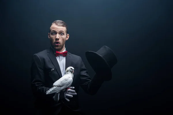 Mago scioccato mostrando trucco con colomba e cappello in camera oscura con fumo — Foto stock