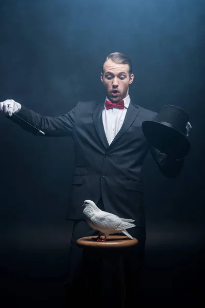Schockierter Zauberer zeigt Trick mit Taube, Zauberstab und Hut in dunklem Raum mit Rauch — Stockfoto