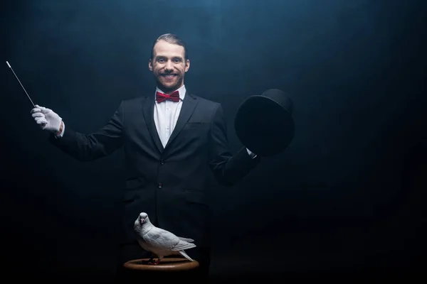 Happy magician showing trick with dove, wand and hat in dark room with smoke — Stock Photo