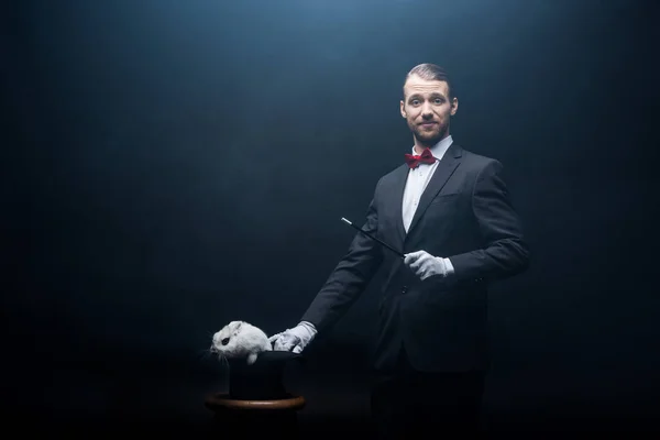 Heureux magicien en costume montrant tour avec baguette et lapin blanc dans le chapeau, chambre sombre avec fumée — Photo de stock