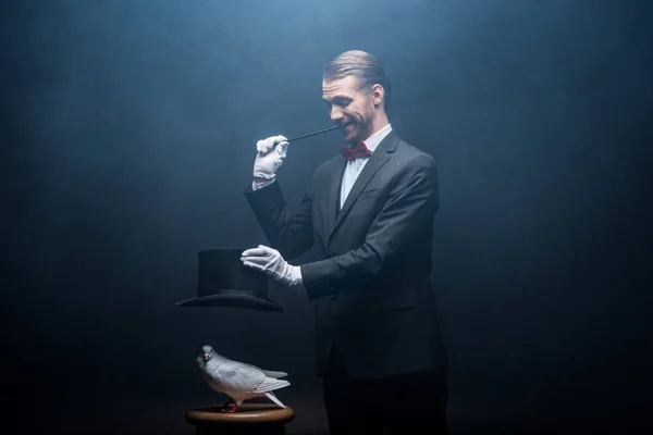 Fröhlicher Zauberer zeigt Trick mit Taube, Zauberstab und Hut in dunklem Raum mit Rauch — Stockfoto