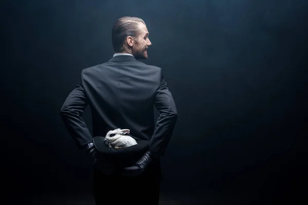 Back view of smiling magician holding white rabbit in hat, dark room with smoke — Stock Photo