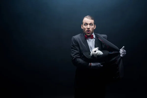 Shocked magician holding white rabbit in hat, dark room with smoke — Stock Photo