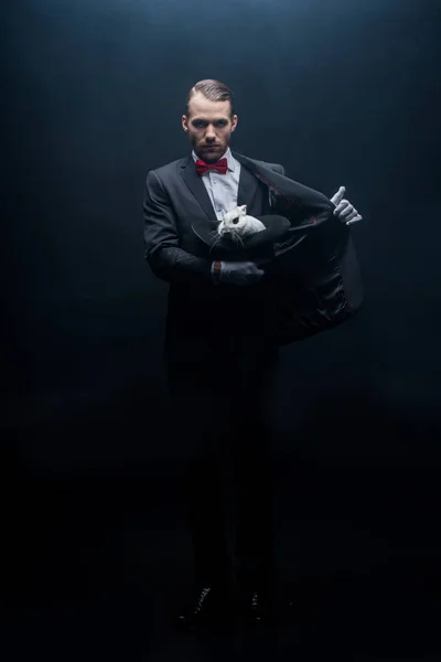 Professional magician in suit holding white rabbit in hat, dark room with smoke — Stock Photo