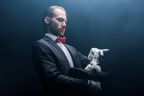 Професійний фокусник бере білого кролика з капелюха, темна кімната з димом — стокове фото