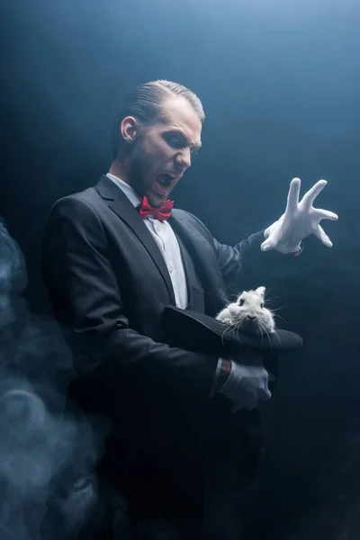 Mago profesional gritando y haciendo gestos sobre conejo blanco en sombrero, cuarto oscuro con humo - foto de stock