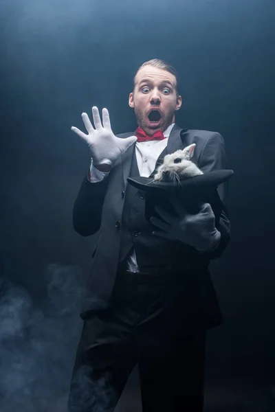 Shocked magician with open mouth holding white rabbit in hat, dark room with smoke — Stock Photo